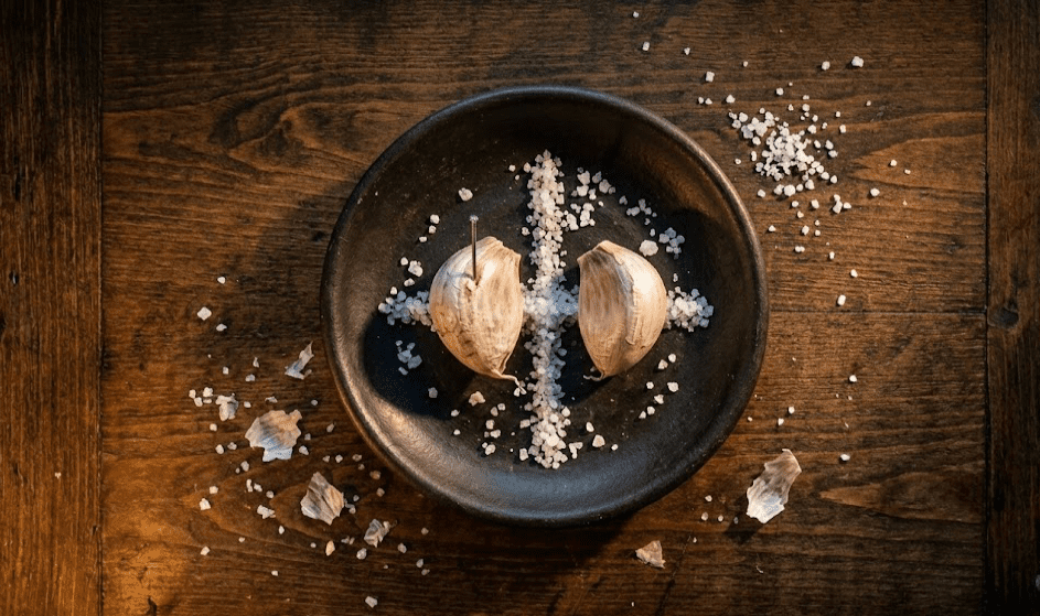 Overhead close-up of two peeled garlic cloves side by side on a dark plate with a pin inserted in one and white salt scattered around them, candlelight from one side