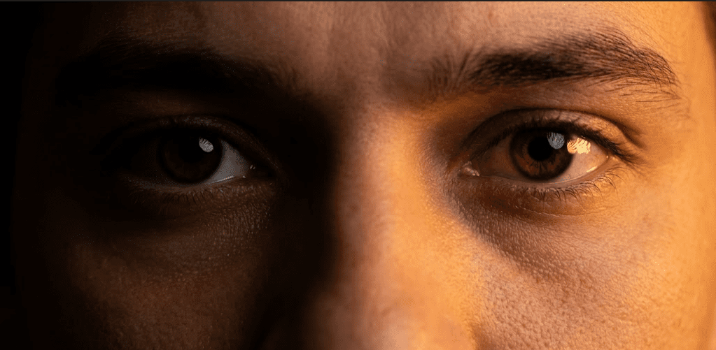 Extreme close-up of a pair of dark human eyes staring directly into the camera, intense and unblinking, amber side lighting, black background