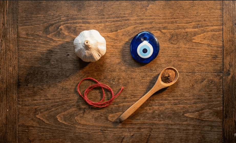 Overhead flat lay of a blue nazar amulet, a garlic clove, a red thread, and ground cinnamon arranged on dark aged wood with amber side lighting