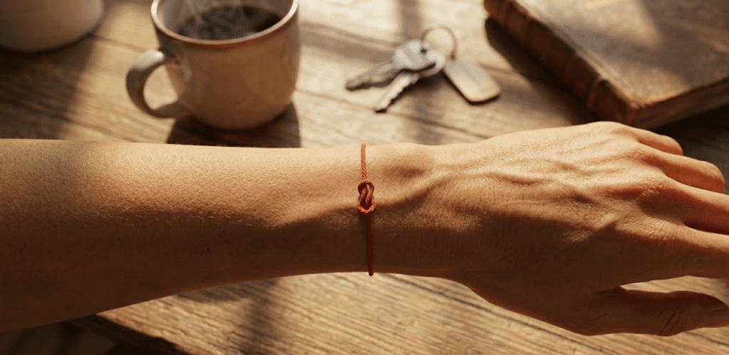 Extreme close-up of a slender wrist with a simple red thread tied around it, warm skin tone, dark background, soft amber light