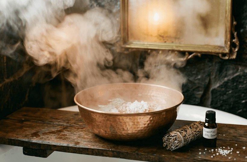 A copper bowl filled with steaming water and coarse sea salt crystals, placed on a dark wooden bath shelf, thick steam rising and catching the light from two beeswax candles in brass holders on either side, a dark stone wall and antique mirror with brass frame visible in the background, a bundle of dried sage and a small glass bottle of essential oil beside the bowl, photorealistic, 35mm film grain, warm humid atmosphere, no people, no faces, focus on the salt crystals and steam texture, ritual cleansing atmosphere, 16:9 aspect ratio
