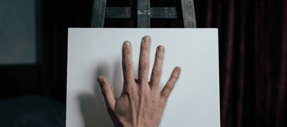 A human hand extended palm-forward in front of a plain white cardboard sheet mounted on a dark wooden easel, the hand is lit by a single beeswax candle in a brass holder from the left, a faint silver-white outline barely visible along the finger edges against the white background, dark bedroom corner with heavy curtains behind the easel, photorealistic, 35mm film grain, extreme shallow depth of field on the hand, warm golden candlelight mixing with cold shadow, no face, no body, only the hand and the white card, subtle etheric layer visible as thin shimmer, 16:9 aspect ratio
