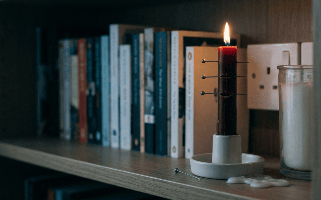 A red dinner candle on a bookshelf with three pins pushed into the wax and one already fallen beside it